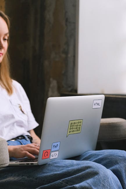 A young woman with long brown hair, wearing a white top, is sitting outdoors on a gray lounge chair with her legs crossed, working on a silver laptop that is decorated with colorful stickers, includin